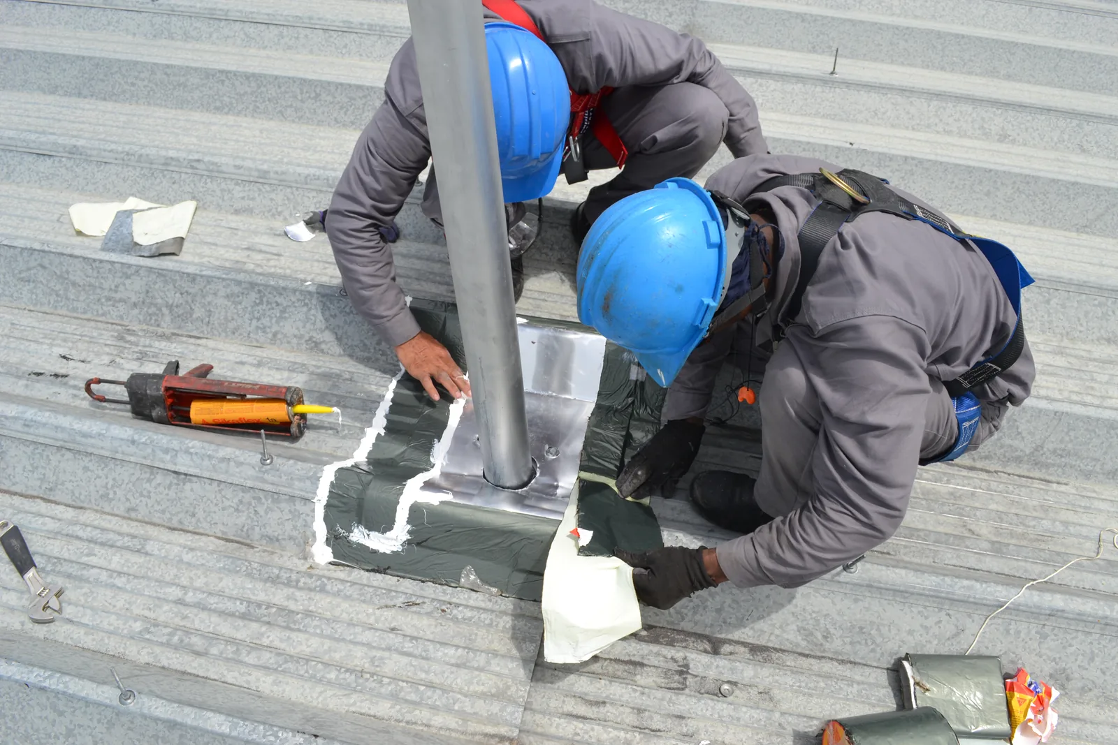 Dos técnicos con arnés de cuerpo completo instalando y sellando la base de un poste de línea de vida sobre cubierta metálica