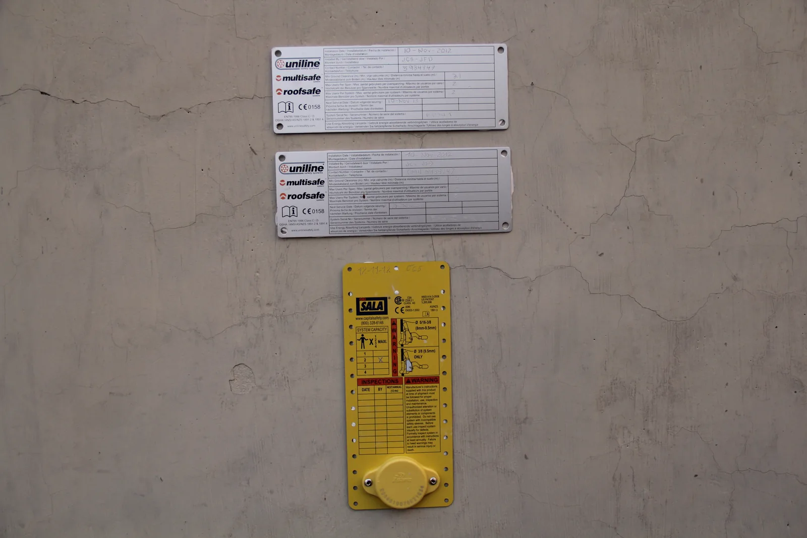 Dos placas de sistema de línea de vida y una placa de registro de inspecciones instaladas sobre un muro de hormigón